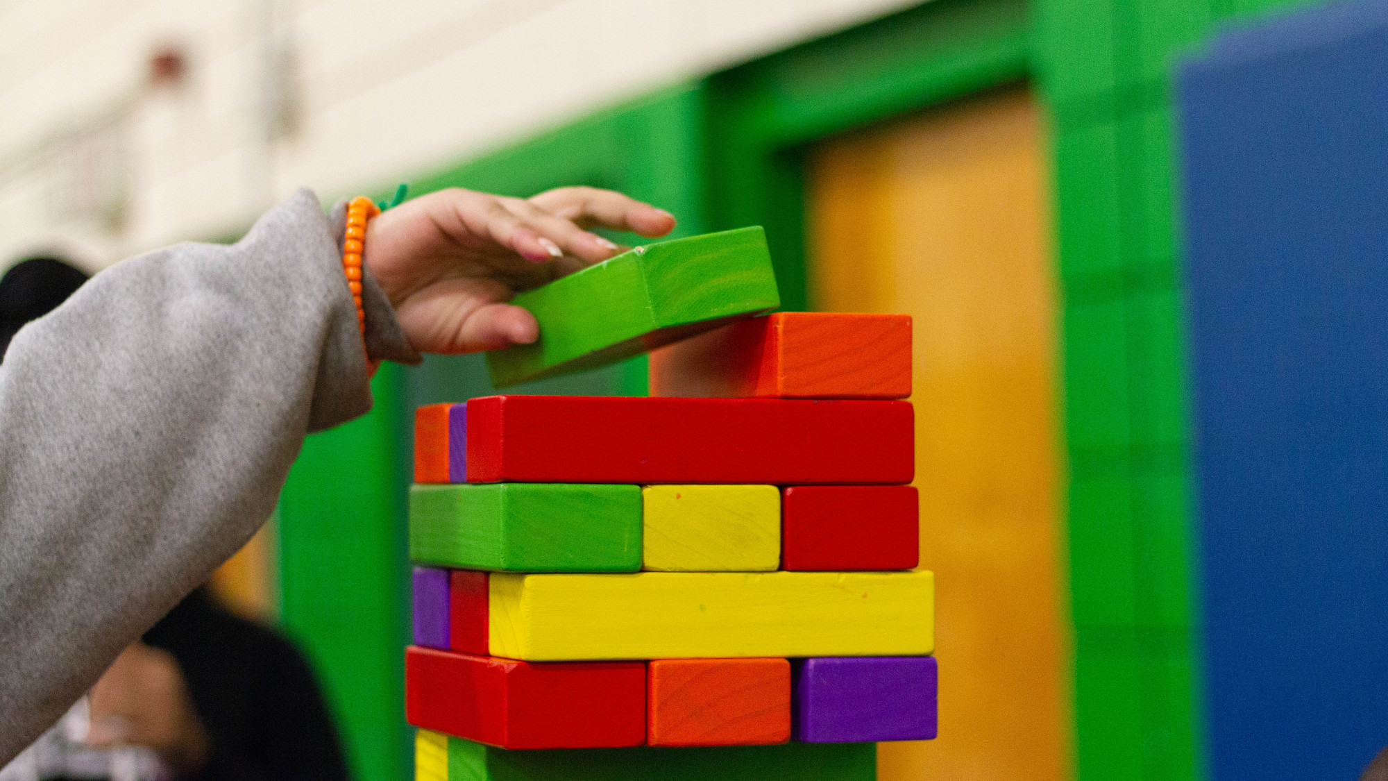 A kids hand puts a green wooden block on a tower built from other wooden block of different colors